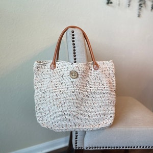 May include: A white crocheted tote bag with brown leather handles and a metal button closure. The bag is sitting on a light gray chair.