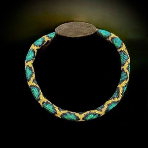 K&ouml;nnte beinhalten: Eine Perlenkette mit geometrischem Muster. Die Halskette weist ein sich wiederholendes Design aus t&uuml;rkis- und goldfarbenen Perlen auf, wodurch ein strukturiertes Aussehen entsteht. Die Halskette wird auf einem dunkelgrauen St&auml;nder vor schwarzem Hintergrund pr&auml;sentiert.