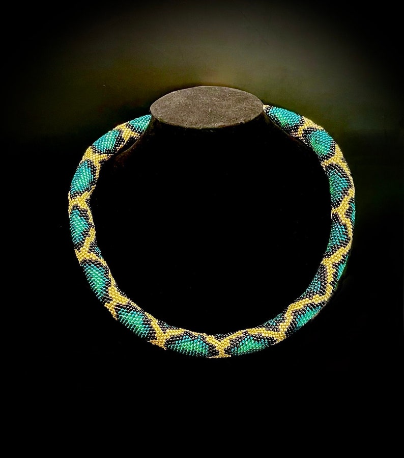 K&ouml;nnte beinhalten: Eine Perlenkette mit geometrischem Muster. Die Halskette weist ein sich wiederholendes Design aus t&uuml;rkis- und goldfarbenen Perlen auf, wodurch ein strukturiertes Aussehen entsteht. Die Halskette wird auf einem dunkelgrauen St&auml;nder vor schwarzem Hintergrund pr&auml;sentiert.