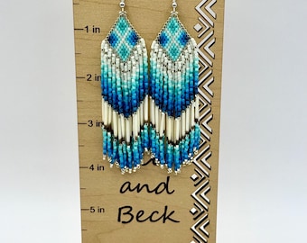 May include: Handmade beaded earrings in shades of blue, turquoise, and white, displayed on a wooden stand. The earrings feature a geometric design at the top and a fringe of beads and silver tubes. The stand has the text "and Beck" and is approximately 5 inches tall.