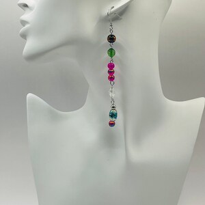 May include: Long silver dangle earrings with colorful beads. The beads are pink, green, brown, and blue. The earrings are hanging from a mannequin's ear.