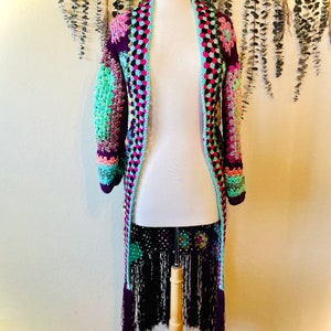 May include: A long, crocheted cardigan with a colorful patchwork design and fringe trim. The cardigan is made with a variety of colors, including purple, pink, green, and black.