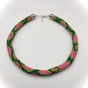 May include: A pink and green beaded necklace with a silver clasp. The necklace is made of small beads that are woven together to create a snake-like pattern.