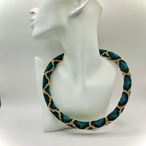 K&ouml;nnte beinhalten: Halskette mit einem geometrischen Muster in Teal und Gold. Die Halskette wird auf einer wei&szlig;en B&uuml;ste pr&auml;sentiert. Die Perlen erzeugen ein strukturiertes, auff&auml;lliges Design, perfekt, um jedem Outfit einen Farbtupfer zu verleihen.