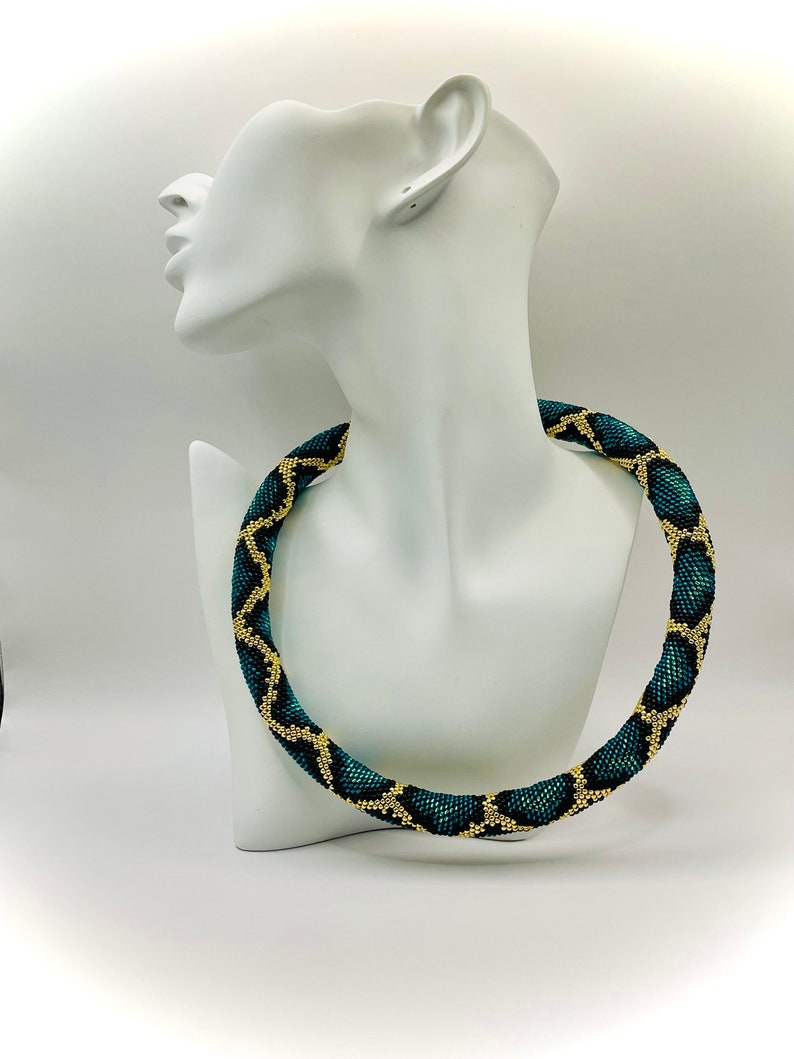 K&ouml;nnte beinhalten: Halskette mit einem geometrischen Muster in Teal und Gold. Die Halskette wird auf einer wei&szlig;en B&uuml;ste pr&auml;sentiert. Die Perlen erzeugen ein strukturiertes, auff&auml;lliges Design, perfekt, um jedem Outfit einen Farbtupfer zu verleihen.