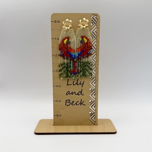 May include: Beaded earrings with a parrot design in red, blue, and green. The earrings are topped with gold-colored flower studs. The earrings are displayed on a wooden stand that reads "Lily and Beck".
