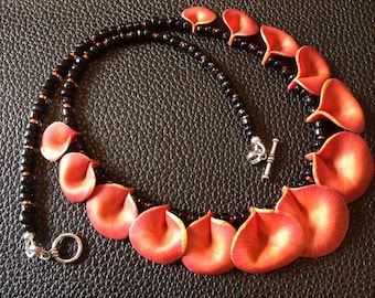 May include: A handcrafted necklace featuring a series of red and orange petal-shaped beads. The petals are interspersed with black beads and connected by a black beaded strand. The necklace is approximately 18 inches long.