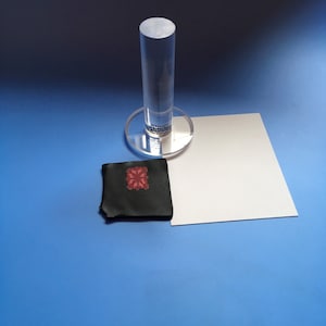 Peut inclure: Un cylindre et une base circulaire en acrylique transparents avec du texte, une carte rectangulaire blanche et un petit carré noir avec un motif floral rouge sur une surface bleue.
