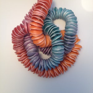 May include: Abstract art piece composed of multiple, interconnected, circular forms. The forms are made of layered, petal-like elements in shades of orange, pink, purple, and teal, creating a textured, three-dimensional effect against a white background.
