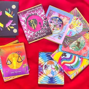 May include: A collection of colorful oracle cards with various illustrations and text. The cards feature images of scales, rainbows, and abstract designs. The cards are arranged on a red surface.