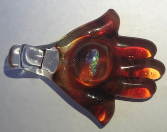 May include: A glass hand charm with a clear glass loop for hanging. The hand is a deep red color with a clear glass center that has an iridescent opal inside.
