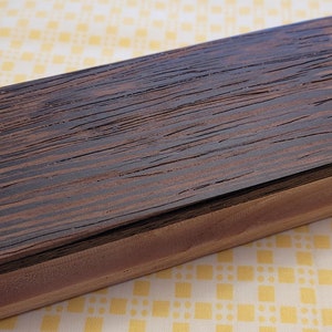 Op de afbeelding: Een ovale houten doos met een donkerbruin, gestreept deksel en een lichtere bruine basis. De doos heeft een natuurlijk houtnerfpatroon. Het deksel staat iets open en onthult de binnenkant.