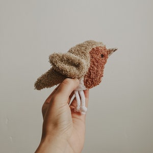 Puede incluir: Un pájaro de peluche hecho de tela marrón, beige y blanca. El pájaro tiene un pequeño pico y una cuerda blanca que cuelga de sus pies.