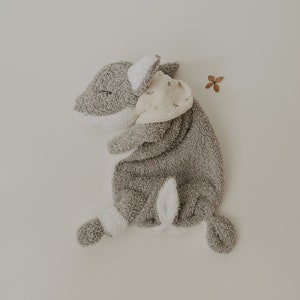 May include: A gray plush toy shaped like a fox with white accents. The toy is lying on its side with its head resting on its paws. The toy has a small, light pink bandana with a floral pattern around its neck.