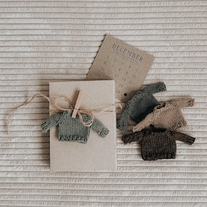 May include: A neutral-toned gift box is adorned with a miniature green sweater, secured with twine and a wooden clip. Several other tiny sweaters in shades of green, brown, and tan are scattered nearby, along with a December calendar.