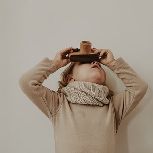 Könnte beinhalten: Ein Kind trägt einen beigefarbenen Pullover und einen Strick Schal und hält eine hölzerne Spielzeugkamera vor seine Augen. Die Kamera ist aus hellem braunem Holz mit einem dunkelbraunen Objektiv.