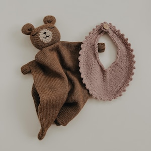 May include: A brown knitted bear comforter with a cream face and closed eyes. A matching pink bib with a scalloped edge and a wooden button closure is next to the comforter. The items are made of soft, knitted material.