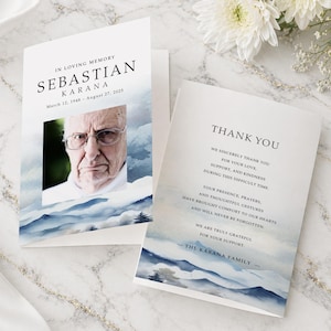 May include: A folded funeral memorial card featuring a portrait of an elderly person with glasses, the text IN LOVING MEMORY SEBASTIAN KARANA March 12, 1948 - August 27, 2025, and a matching thank you card with blue watercolour mountain landscape art.