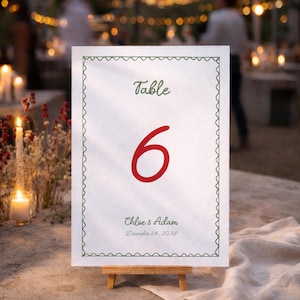 May include: A white table number card with a green scalloped border. The word "Table" is written in green script above a large red number "6". Below, the names "Chloe & Adam" and the date "December 24, 2038" are printed in green.