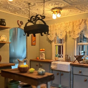 Op de afbeelding: Een miniatuurkeuken met een witte porseleinen gootsteen, houten kasten en een hangende potrek. De keuken heeft een decoratief haanbeeldje, een weegschaal met appels en een kan op een tafel.