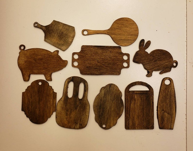 May include: A set of 12 wooden cutouts in various shapes, including a pig, a rabbit, a spatula, and a variety of kitchen utensils. The cutouts are all made of dark brown wood and have a rustic, hand-crafted look.