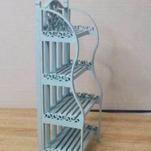 Puede incluir: Una estantería en miniatura de color azul claro con tres niveles y adornos decorativos. La estantería tiene un diseño curvo y soportes verticales, con listones horizontales para cada nivel. La parte superior presenta un diseño floral ornamentado.