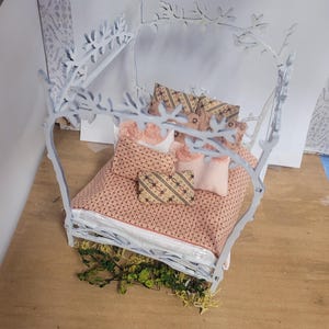 May include: A miniature white bed frame with a canopy, adorned with floral and leaf cutouts. The bed is made up with a pink patterned comforter and pillows. Greenery is placed at the base of the bed.