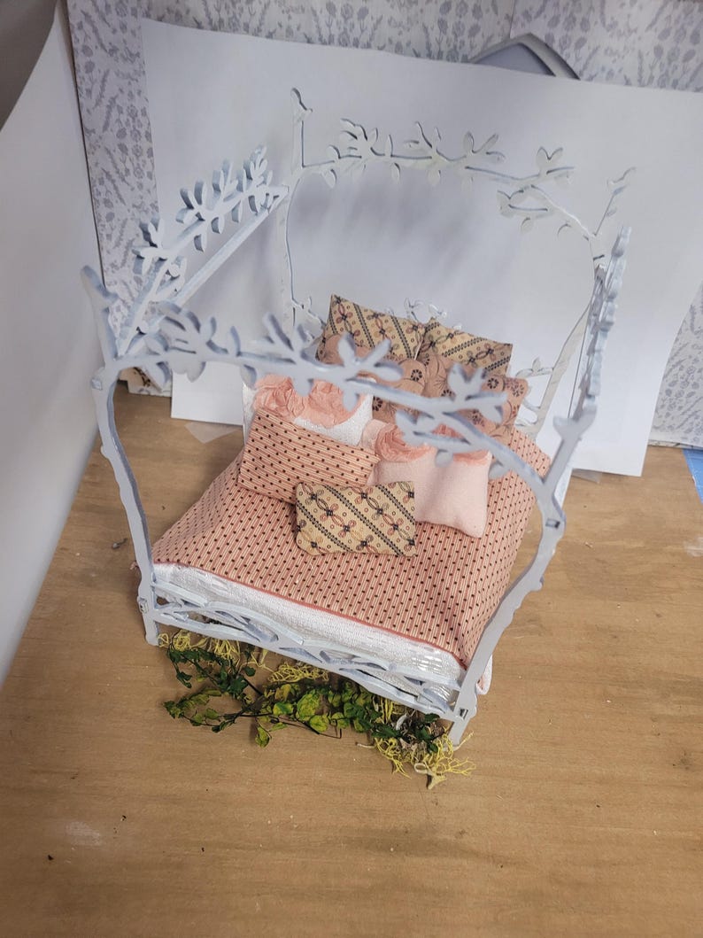 May include: A miniature white bed frame with a canopy, adorned with floral and leaf cutouts. The bed is made up with a pink patterned comforter and pillows. Greenery is placed at the base of the bed.