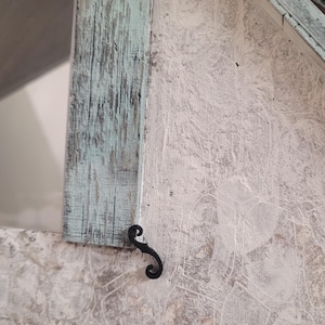 Op de afbeelding: Close-up van een decoratieve houten hoek van een huis. Het huis heeft verweerd hout in tinten lichtblauw en wit. Een decoratieve zwarte metalen sluiting is aan het huis bevestigd.