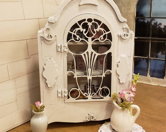 May include: White painted wooden cabinet with a glass door and shelves. The cabinet has a decorative arched top and intricate metalwork on the door. Two small white vases with pink flowers are on either side of the cabinet.