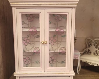 May include: A white painted miniature cabinet with glass doors and a drawer. The cabinet has a decorative top and floral accents on the glass. The cabinet is painted in a distressed style.