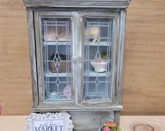 May include: A distressed white and blue cabinet with glass doors and a decorative metal top. The cabinet has a sign that reads "Fresh from the Market" with a list of flowers. A small gray pot with red flowers sits in front of the cabinet.
