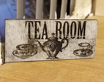 Puede incluir: Un letrero de madera rústico con el texto "TEA ROOM" en letras negras. El letrero presenta una tetera y dos tazas de té con platillos.
