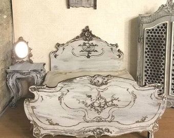 May include: White painted wooden dollhouse bed with ornate carvings and a distressed finish. The bed has a white headboard and footboard with intricate details. The bed is made of wood and has a white finish.