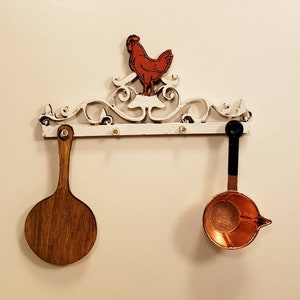 May include: White metal wall mounted rack with a rooster design. The rack has two hooks, one holding a wooden spoon and the other holding a copper ladle.