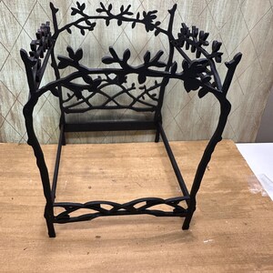 May include: Black metal decorative plant stand with a branch and leaf design. The stand has a rectangular base and a decorative back with branches and leaves. The stand is on a wooden surface.