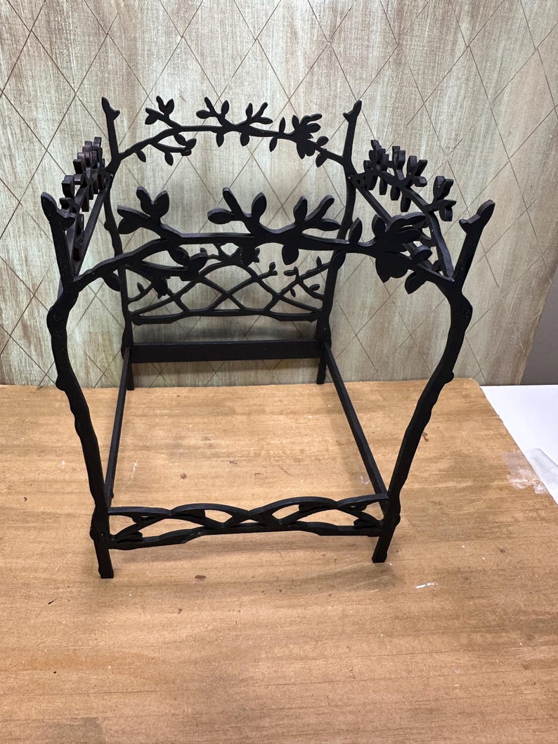 May include: Black metal decorative plant stand with a branch and leaf design. The stand has a rectangular base and a decorative back with branches and leaves. The stand is on a wooden surface.