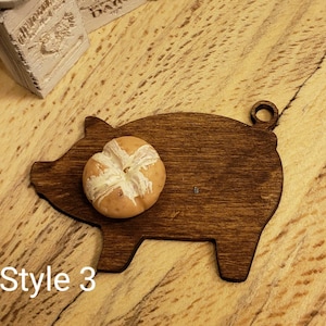 May include: A dark brown wooden pig-shaped cutting board with a round bun on top. The board has a hole in the top for hanging.  Style 3.