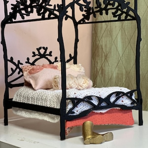 May include: A miniature black metal bed frame with a floral design on the headboard and canopy. The bed is made up with a white knitted blanket, pink pillows, and a coral-colored blanket. A small gold boot sits in front of the bed.