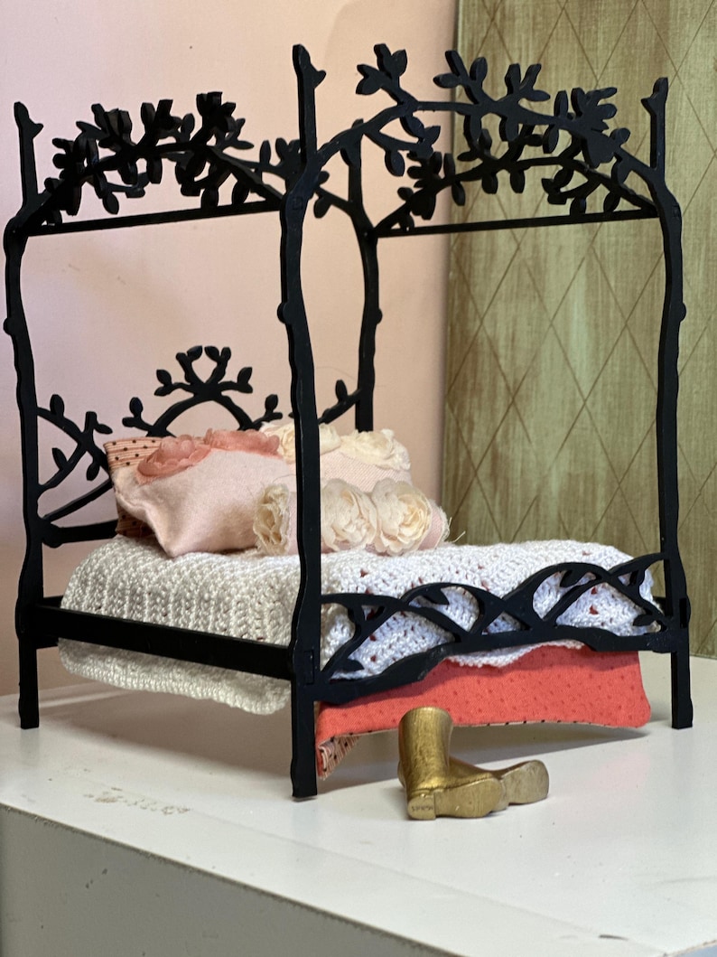 May include: A miniature black metal bed frame with a floral design on the headboard and canopy. The bed is made up with a white knitted blanket, pink pillows, and a coral-colored blanket. A small gold boot sits in front of the bed.