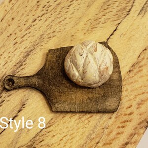 May include: A small, round, white and brown bread loaf on a dark brown wooden cutting board with a handle. The cutting board is on a light brown wooden surface. The text "Style 8" is visible in the lower left corner of the image.