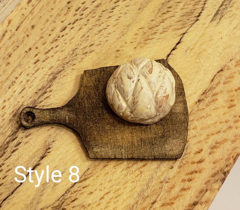 May include: A small, round, white and brown bread loaf on a dark brown wooden cutting board with a handle. The cutting board is on a light brown wooden surface. The text "Style 8" is visible in the lower left corner of the image.