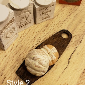 May include: Two miniature loaves of bread on a small wooden cutting board. The bread is white with a light brown crust and a swirl design. The cutting board is dark brown with a rounded handle.