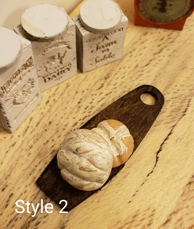 May include: Two miniature loaves of bread on a small wooden cutting board. The bread is white with a light brown crust and a swirl design. The cutting board is dark brown with a rounded handle.
