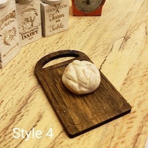 May include: A miniature wooden cutting board with a white bread loaf on top. The cutting board has a dark brown wood grain finish. The bread loaf is slightly oval shaped and has a light brown crust.