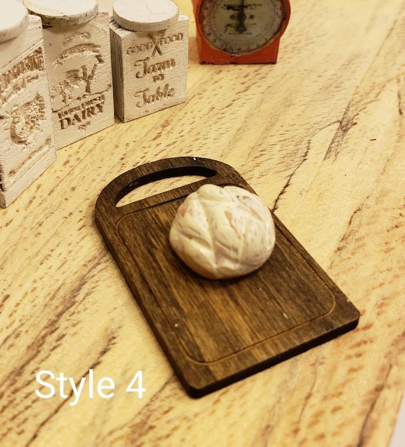 May include: A miniature wooden cutting board with a white bread loaf on top. The cutting board has a dark brown wood grain finish. The bread loaf is slightly oval shaped and has a light brown crust.