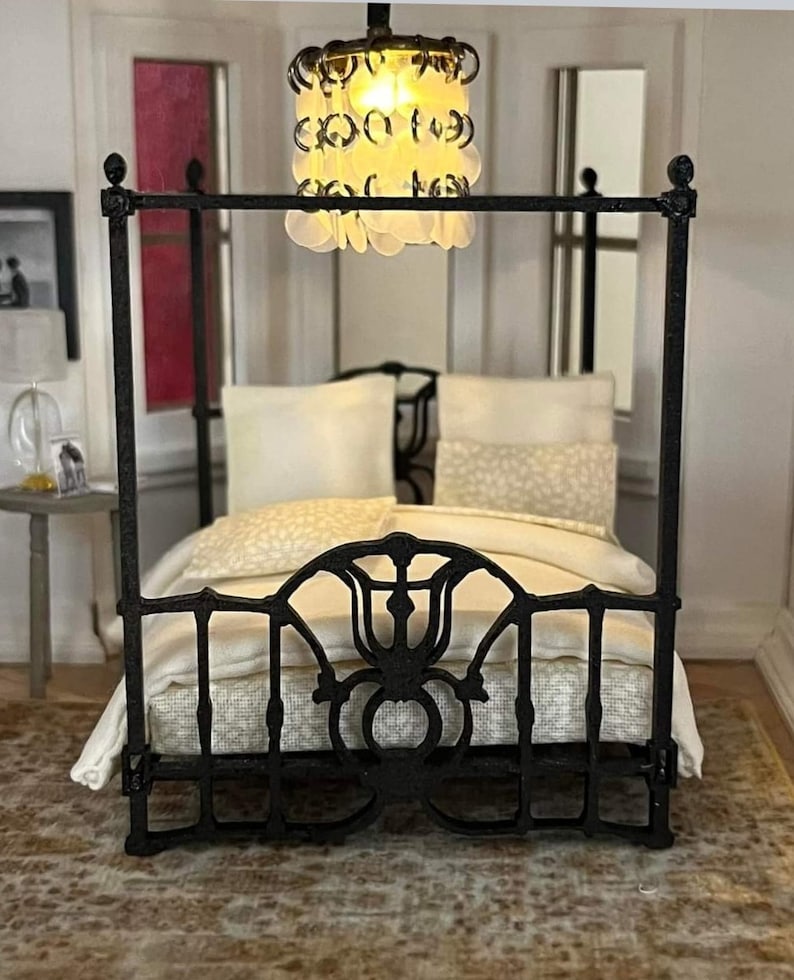 Puede incluir: Una cama con dosel de metal negro con un edred&oacute;n y almohadas blancas. La cama est&aacute; en una habitaci&oacute;n con techo y paredes blancas. Una l&aacute;mpara de ara&ntilde;a con cuentas de vidrio blancas cuelga sobre la cama.