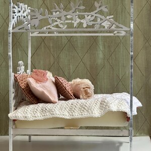 May include: A miniature white bed with a decorative floral headboard and footboard. The bed is adorned with pink and brown pillows, a cream-colored mattress, and a white crocheted blanket. The bed is a dollhouse or decorative item.