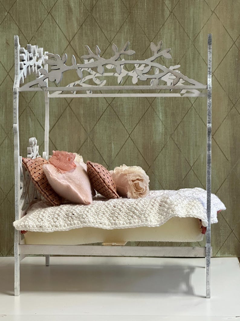 May include: A miniature white bed with a decorative floral headboard and footboard. The bed is adorned with pink and brown pillows, a cream-colored mattress, and a white crocheted blanket. The bed is a dollhouse or decorative item.