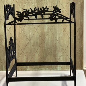 May include: A black metal doll bed with a floral design on the headboard and footboard. The bed frame is rectangular with a horizontal bar for the mattress. The headboard features a decorative floral vine.